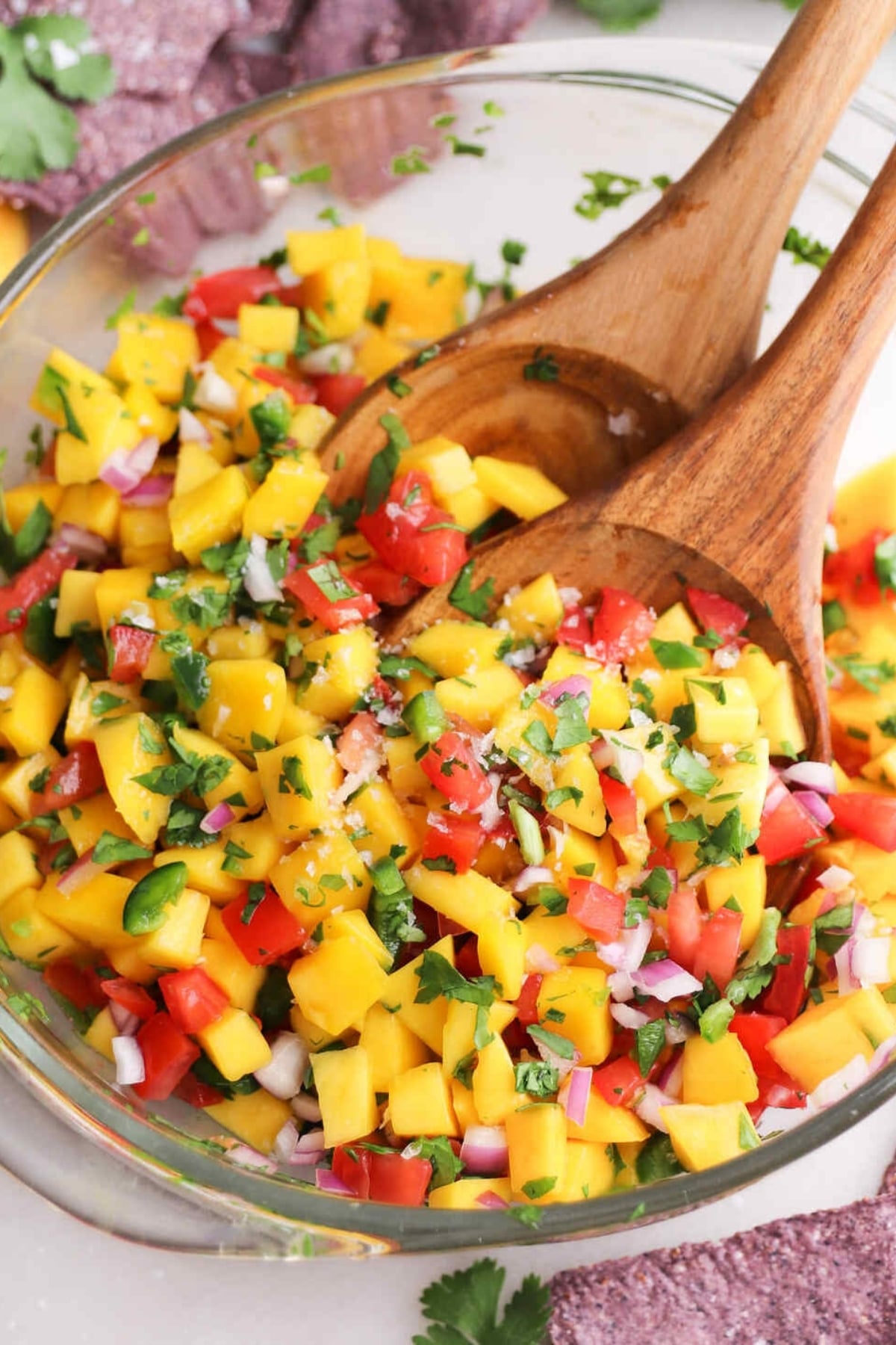 A glass bowl of mango pico de gallo with chopped mango, tomatoes, red onion, cilantro, and jalapeño, mixed with wooden serving utensils—a vibrant salsa fresca recipe perfect for any occasion.