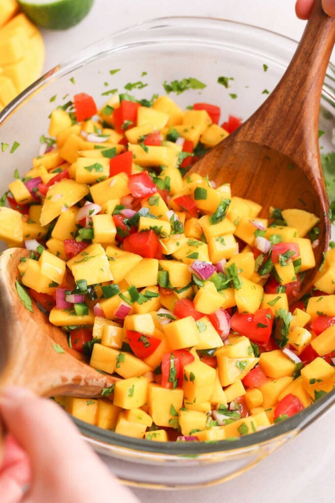 A glass bowl of salsa fresca with chopped mango, red onion, tomato, cilantro, and jalapeño is being mixed with wooden spoons.