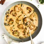A skillet with pork medallions in a creamy mushroom sauce, garnished with herbs. A spoon rests in the pan, and fresh chopped parsley is nearby.