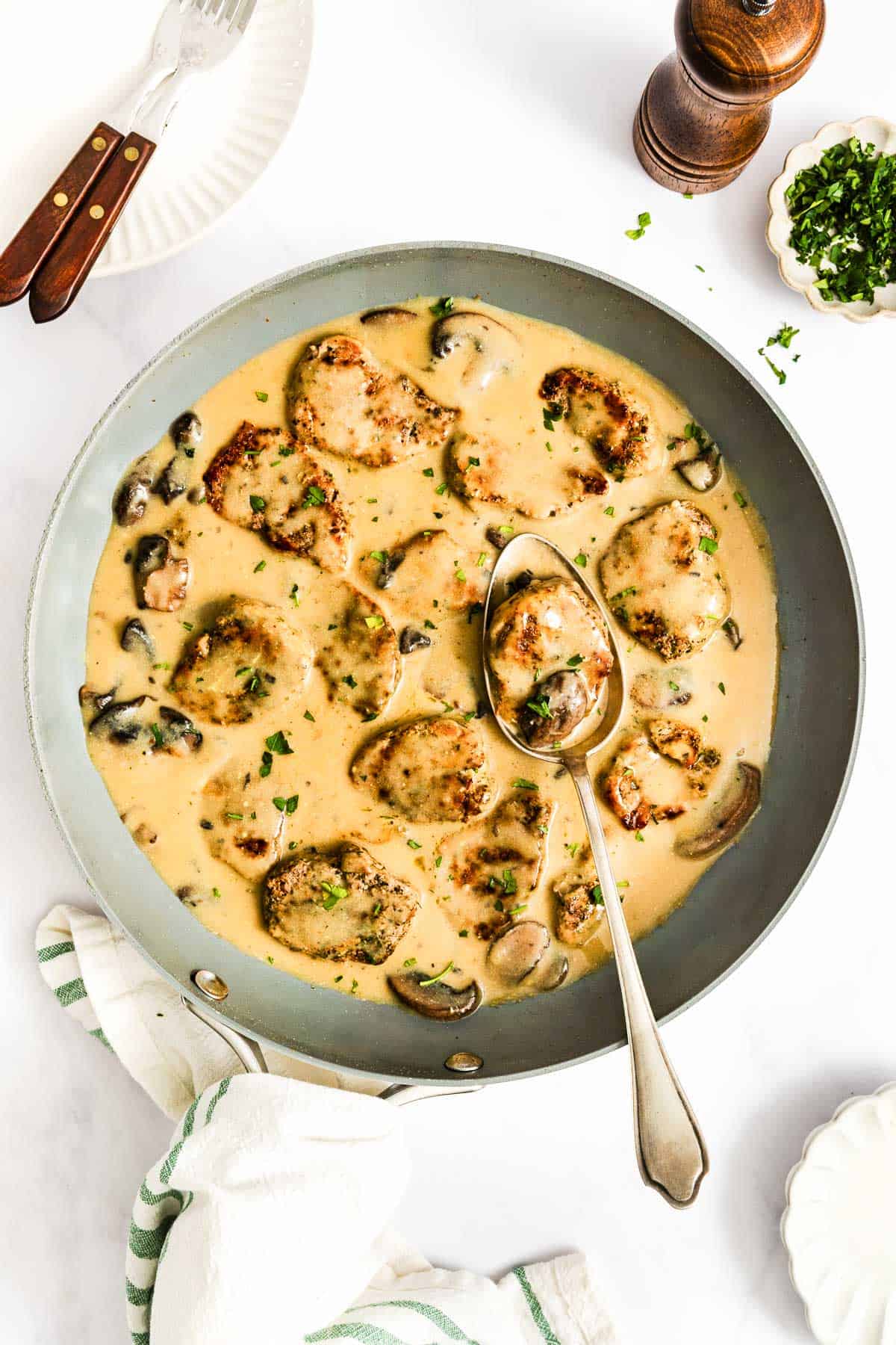 A skillet with chicken medallions in a creamy mushroom sauce, garnished with herbs. A spoon rests in the pan, and fresh chopped parsley is nearby.