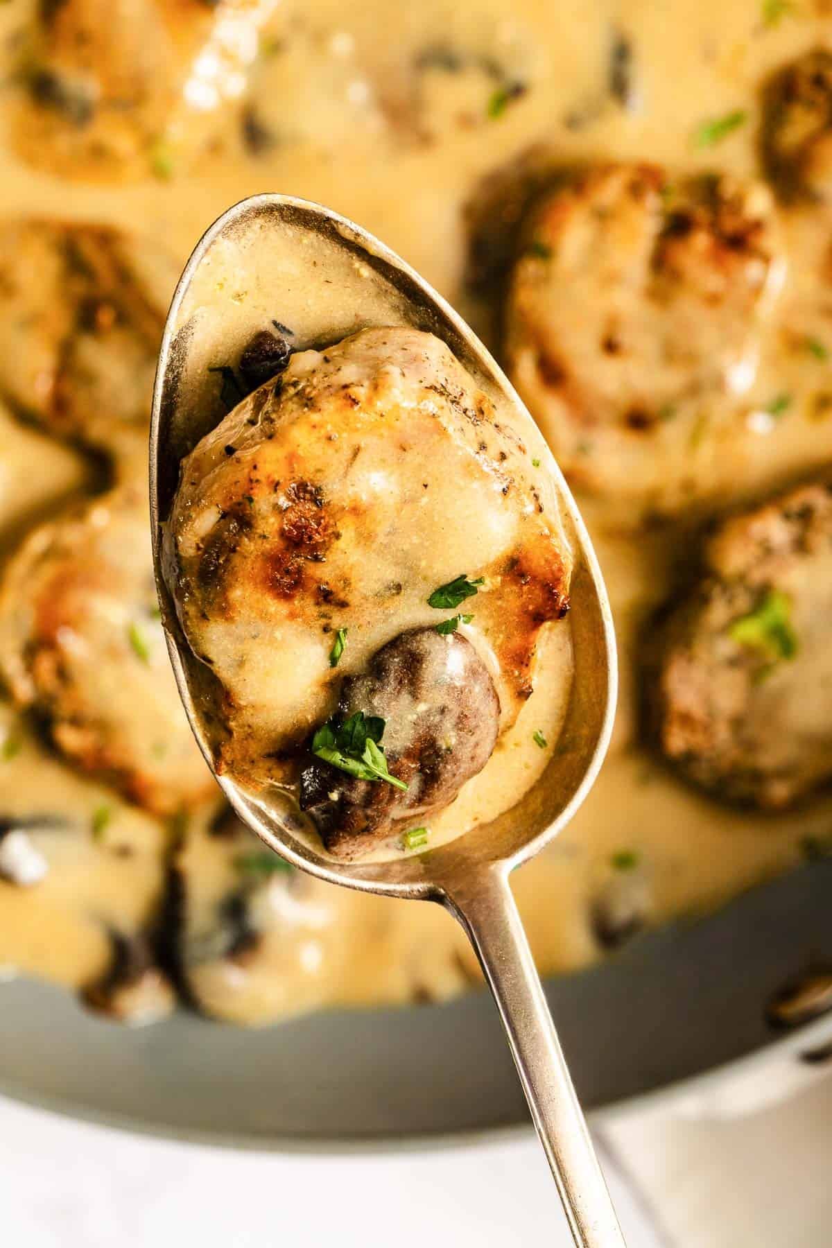 A close-up of a spoon holding a piece of meat with mushrooms in a creamy sauce, with more pieces in the background.