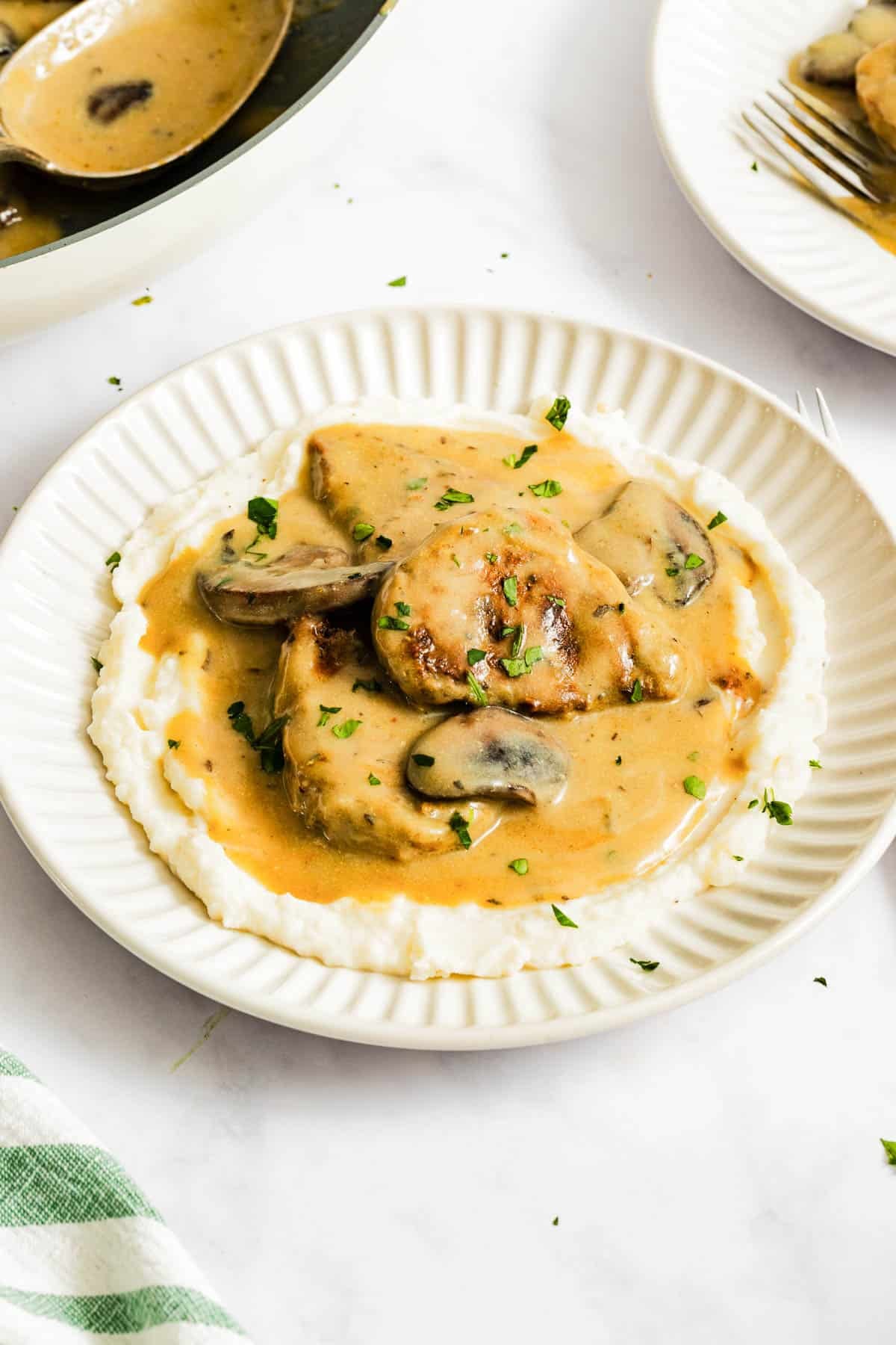 A plate of mashed potatoes topped with pieces of meat and mushrooms in a creamy brown sauce, garnished with chopped herbs.