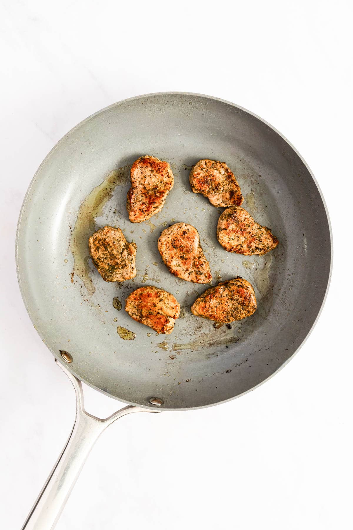 Six seared pieces of meat cooking in a nonstick frying pan on a white background.