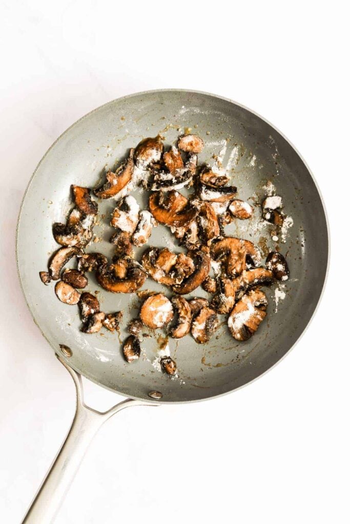 Sliced mushrooms sautéing in a grey frying pan, sprinkled with flour.