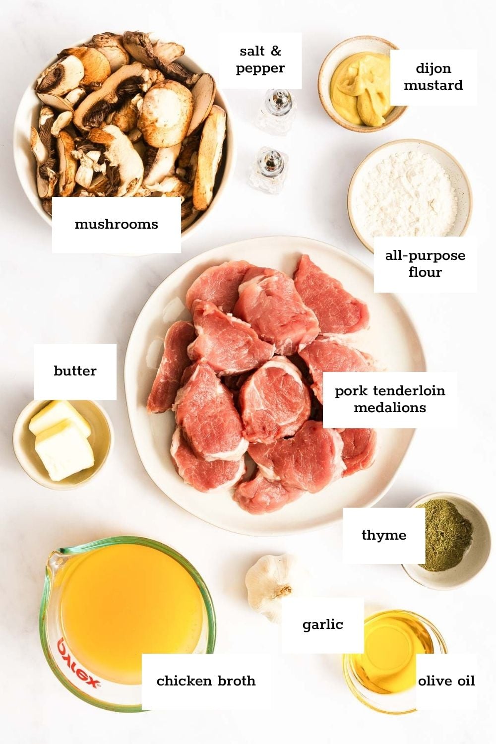 Top-down view of ingredients for a pork dish, including pork medallions, mushrooms, butter, flour, thyme, garlic, olive oil, chicken broth, Dijon mustard, salt, and pepper.