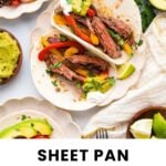 Two steak fajitas with grilled peppers and onions on tortillas, topped with guacamole, are served on white plates with lime wedges and avocado on the side.