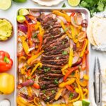 Sheet pan fajitas with cooked sliced steak, red and yellow bell peppers, and red onions, surrounded by tortillas, avocado, lime, guacamole, herbs, and plates with forks.