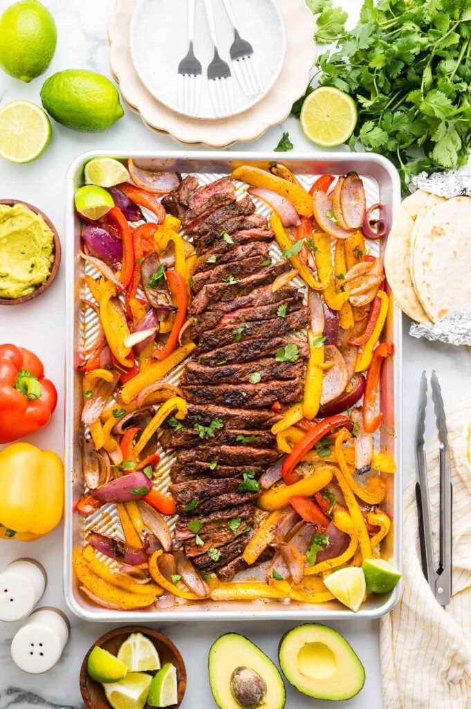 Sheet pan fajitas with cooked sliced steak, red and yellow bell peppers, and red onions, surrounded by tortillas, avocado, lime, guacamole, herbs, and plates with forks.