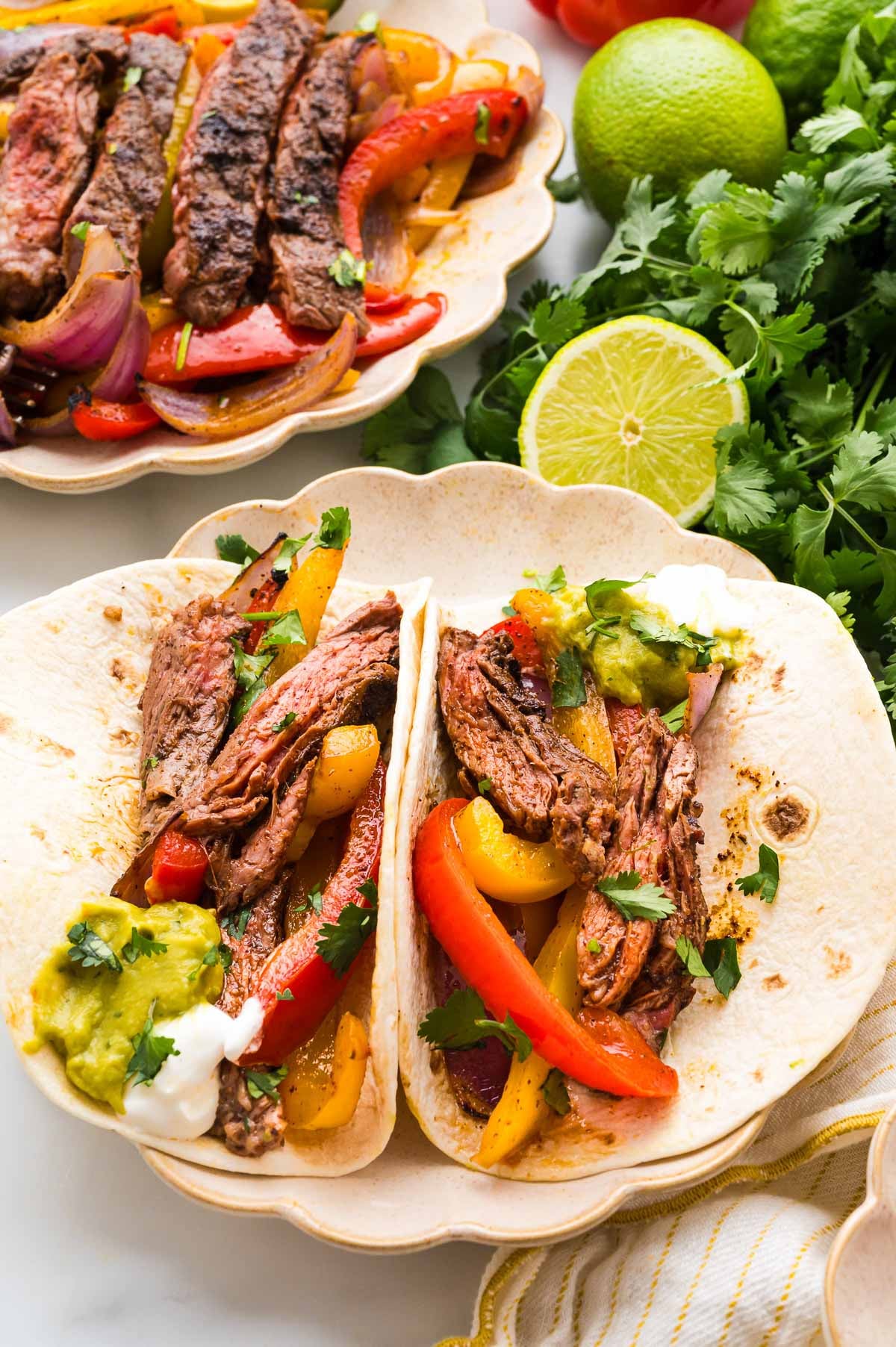 Two flour tortillas filled with grilled steak strips, sautéed bell peppers, onions, guacamole, and sour cream, served on a plate with fresh cilantro and limes nearby.