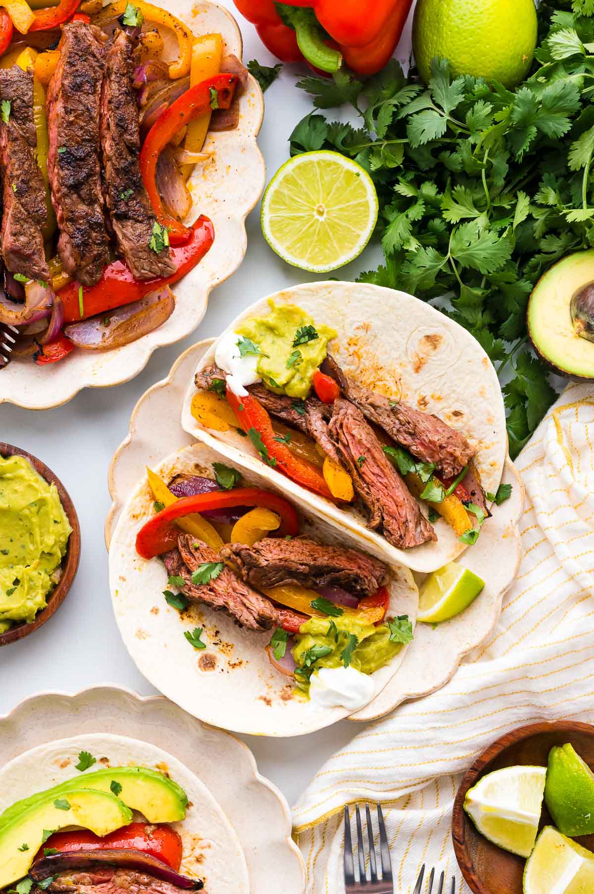 Two beef fajita tacos with grilled vegetables on tortillas, surrounded by sliced steak, guacamole, lime, cilantro, avocado, and bell peppers on a white surface.