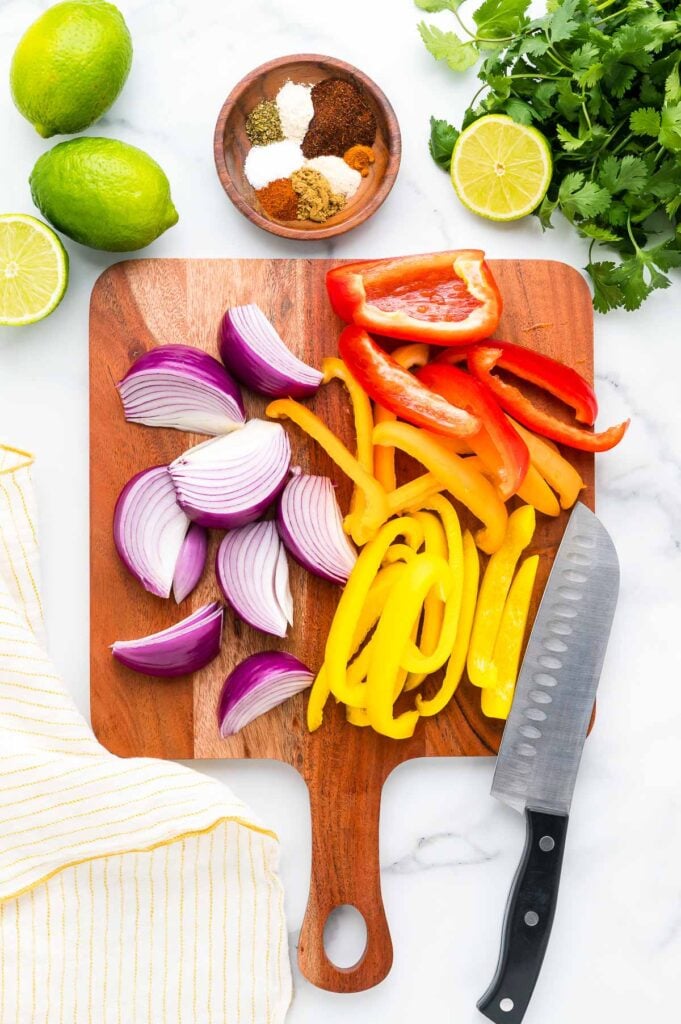 Sliced red onion, red bell pepper, and yellow bell pepper on a wooden cutting board with a knife; limes, cilantro, spices, and a towel nearby.