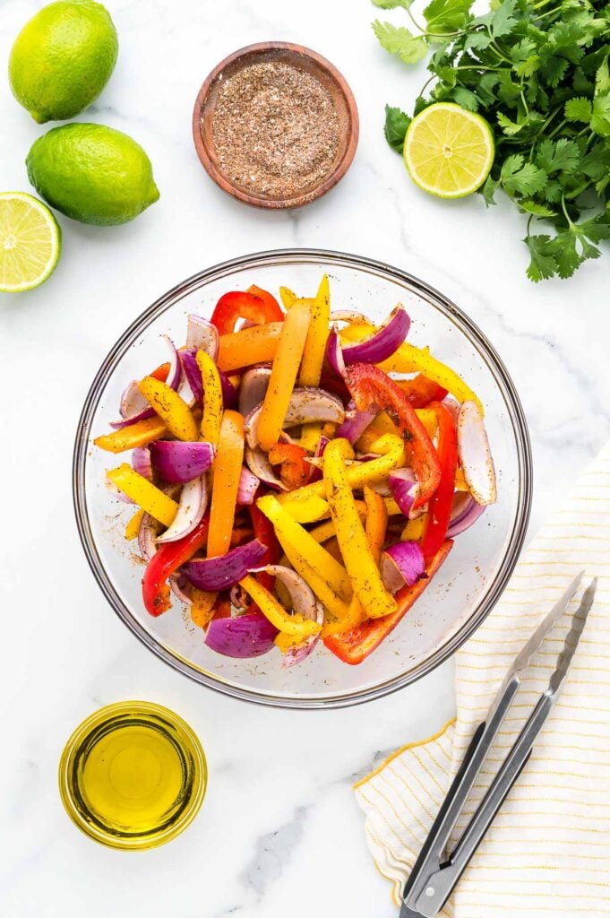 A glass bowl of sliced red and yellow bell peppers and red onions, seasoned with spices. Surrounding items include limes, cilantro, olive oil, a small bowl of spices, and metal tongs.