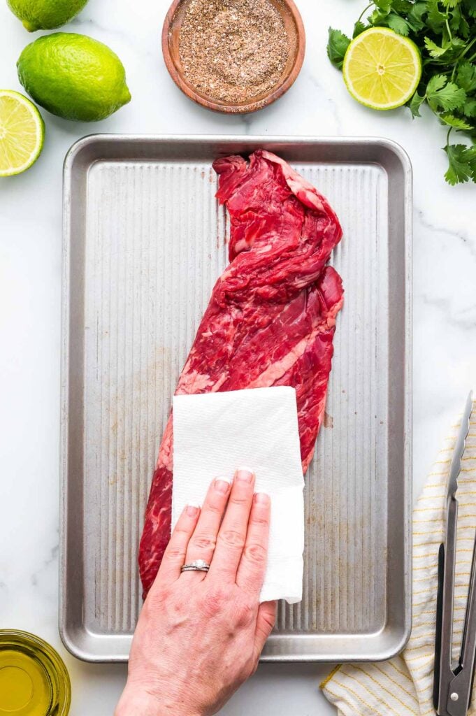 A hand pats a raw skirt steak dry with a paper towel on a metal baking sheet. Limes, seasoning, oil, and cilantro are arranged around the tray.