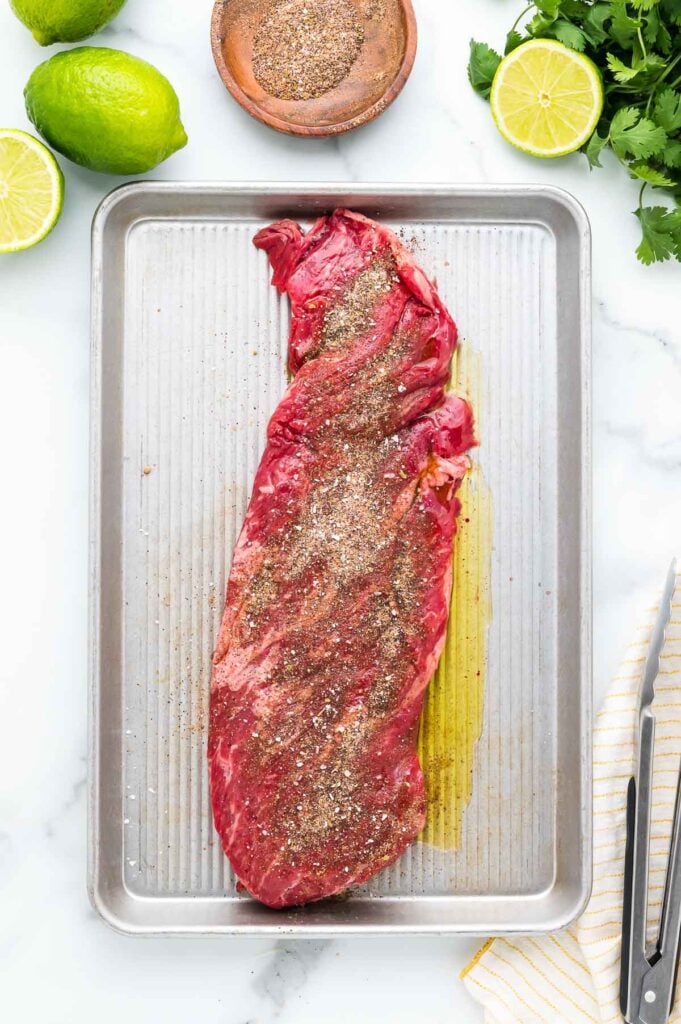 Raw seasoned steak with olive oil and spices on a metal tray, surrounded by lime halves, cilantro, a bowl of seasoning, and utensils.