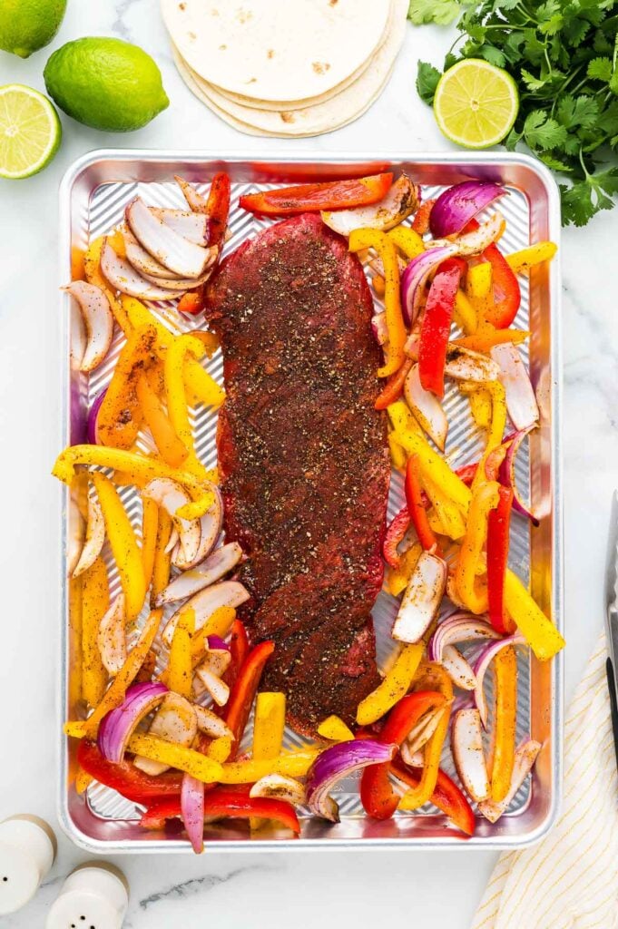 A sheet pan with a seasoned raw steak in the center, surrounded by sliced yellow and red bell peppers and red onion, ready for cooking. Tortillas, limes, and cilantro are nearby.