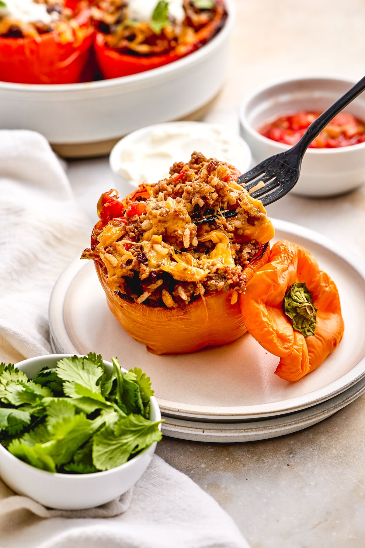 A stuffed orange bell pepper filled with ground meat, rice, and cheese sits on a plate with its top off. A fork holds a portion, and side bowls contain cilantro, sour cream, and diced tomatoes.