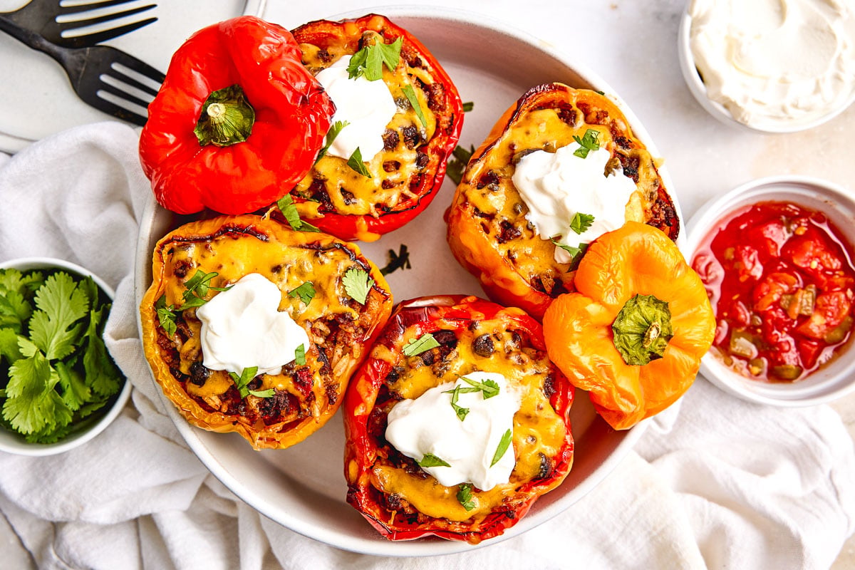 A bowl of stuffed bell peppers topped with melted cheese and sour cream, garnished with cilantro, with sides of salsa, cilantro, and sour cream.