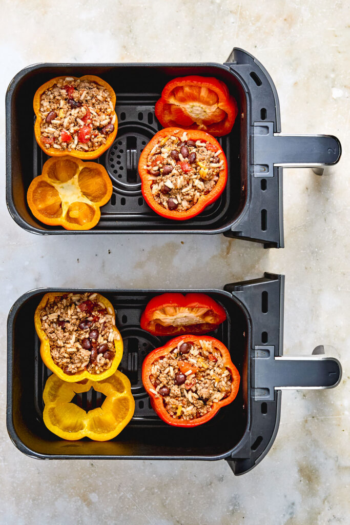 Two air fryer baskets containing halved red and yellow bell peppers stuffed with a ground meat and bean mixture, ready to be cooked.