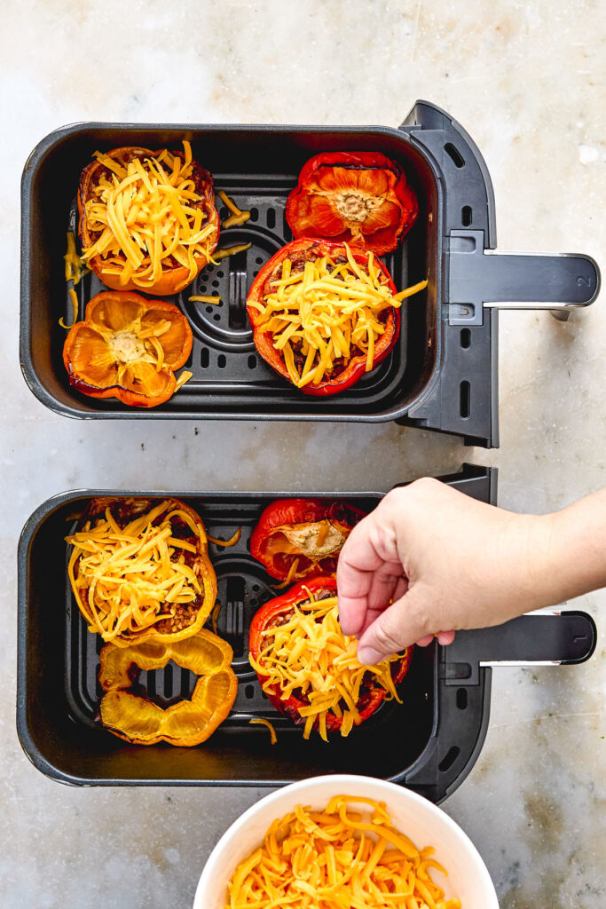 A hand sprinkles shredded cheese onto sliced bell peppers in air fryer baskets, with a bowl of cheese nearby on a light countertop.
