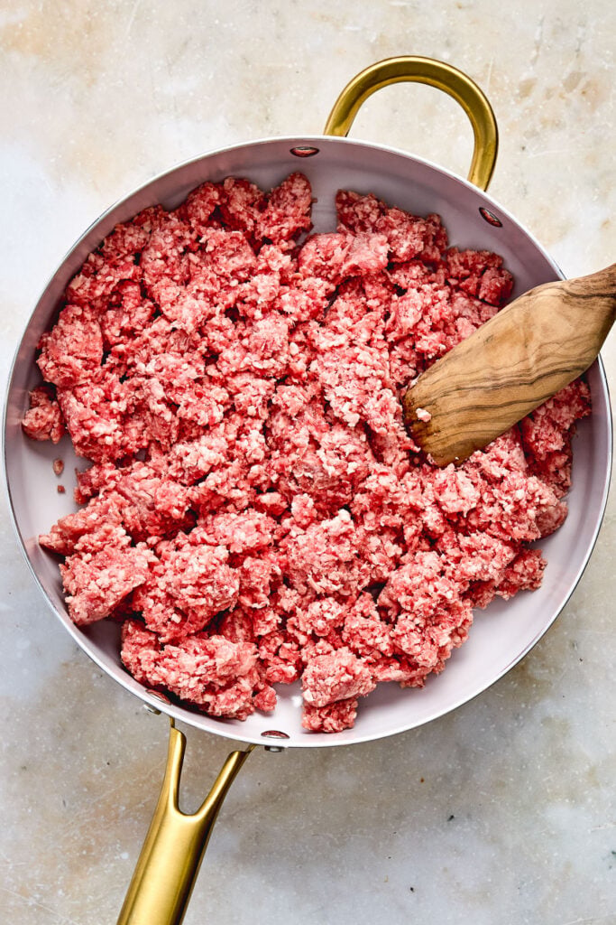 Raw ground beef in a skillet with a wooden spatula on a light-colored surface.