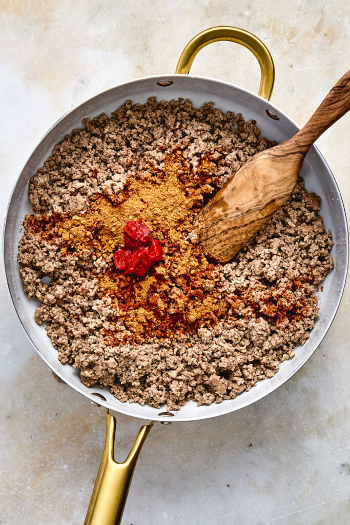 A pan of cooked ground beef with taco seasoning and tomato paste being mixed in with a wooden spatula.