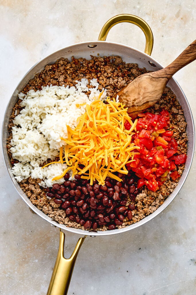 A skillet filled with ground meat, cooked rice, black beans, shredded cheddar cheese, chopped tomatoes, and a wooden spoon.