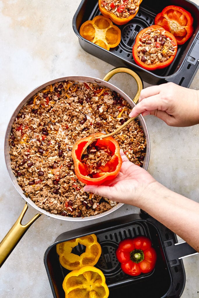 A person stuffs a red bell pepper with a mixture of ground meat, rice, and beans over a pan, with halved peppers and stuffed peppers in air fryer baskets nearby.