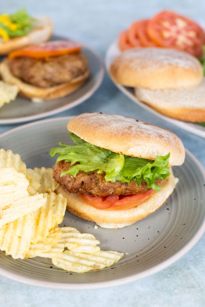 A plated ground turkey hamburger topped with lettuce and tomato, served with potato chips. In the background, more burgers and sandwich toppings are visible.