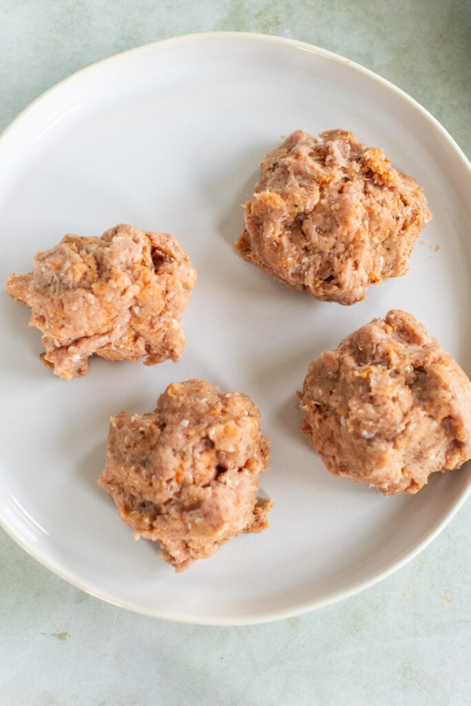 Four raw, unevenly shaped meat mixture portions are arranged on a white plate.