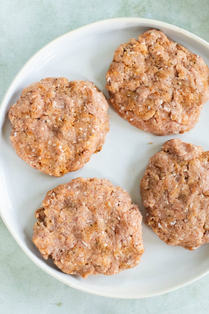 Four uncooked, seasoned ground turkey burger patties are arranged on a white plate against a light background.
