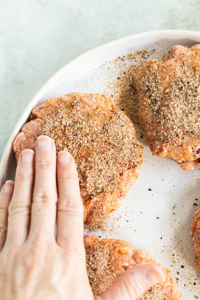 A hand pressing seasoning onto raw turkey burgers arranged in a white dish.