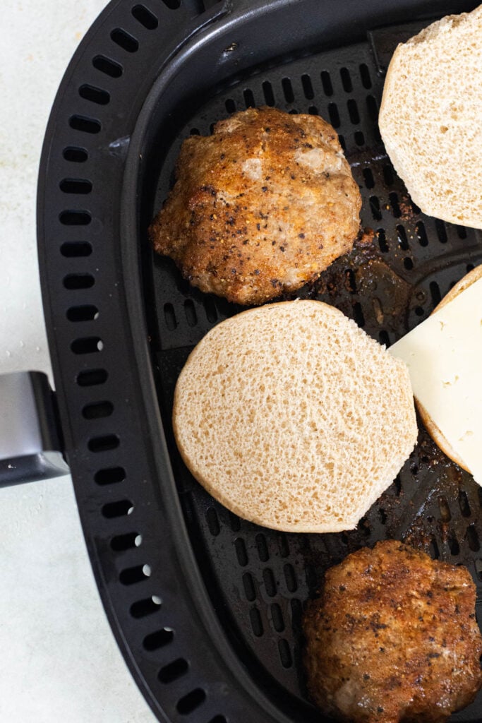 Two cooked burger patties, a sliced burger bun, and a slice of cheese are in the basket of an air fryer.