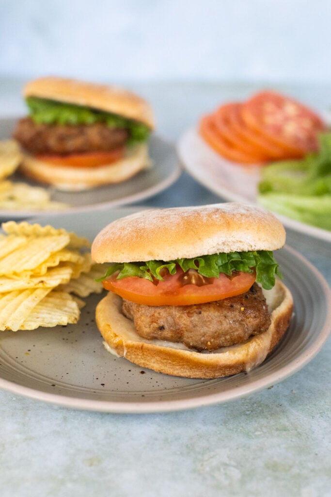 A turkey burger topped with lettuce and tomato next to a pile of potato chips; another burger and sliced vegetables are on a platter in the background.
