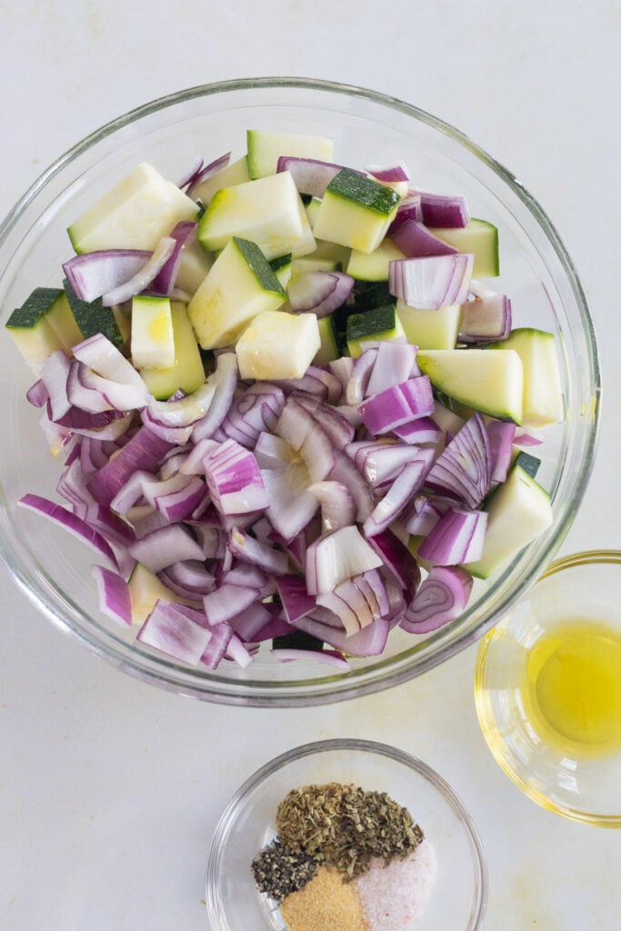 A glass bowl filled with chopped zucchini and red onion, next to a small bowl of olive oil and a dish containing mixed dried herbs, salt, pepper, and garlic powder.