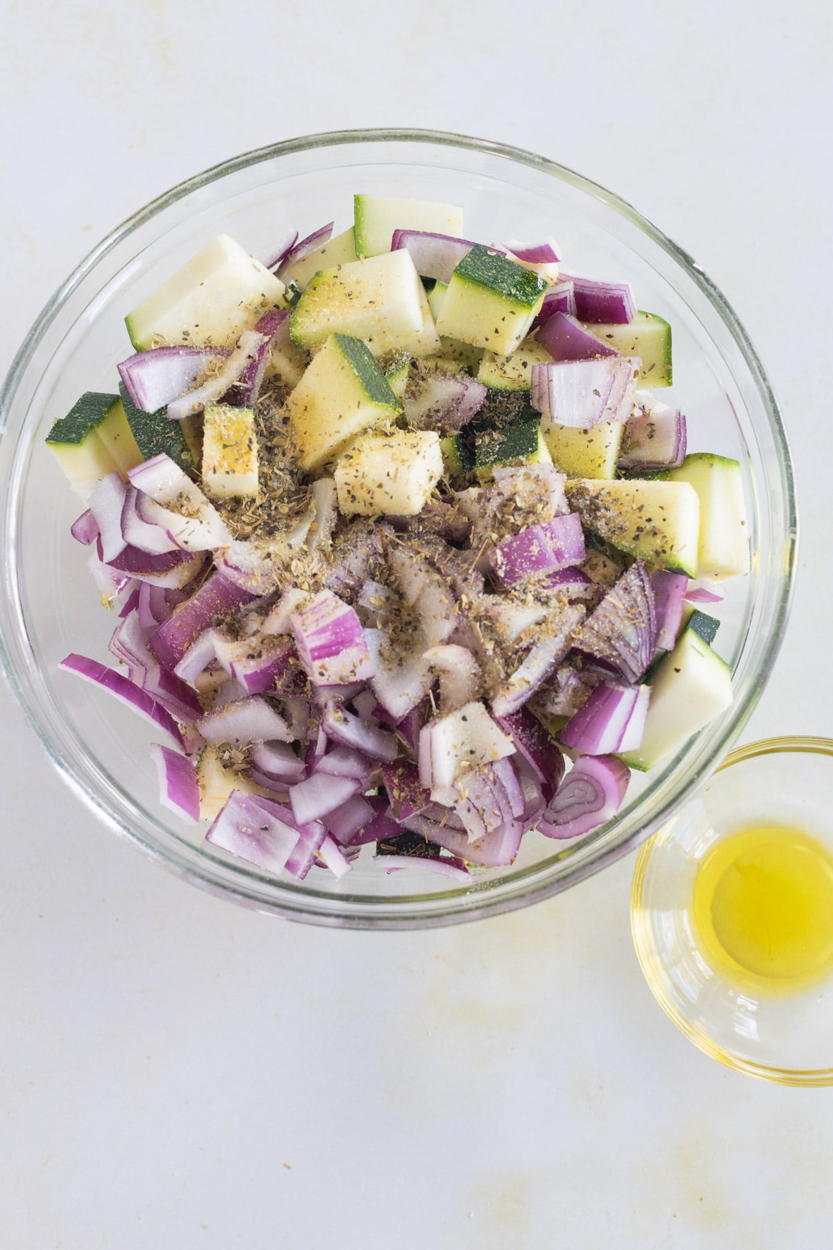 A glass bowl with chopped red onion and zucchini mixed with herbs, next to a small bowl containing olive oil on a white surface.