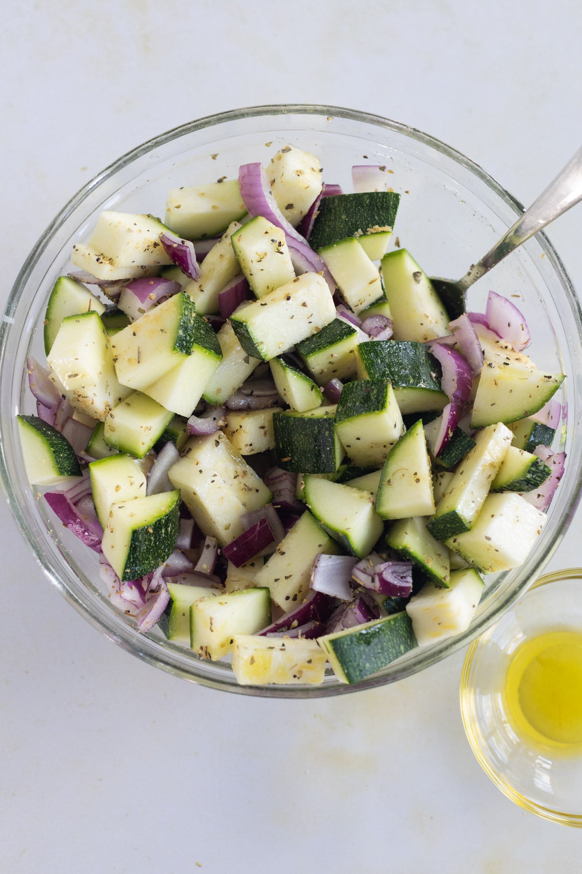 A glass bowl filled with chopped zucchini and red onion, seasoned with herbs, next to a small dish of olive oil.