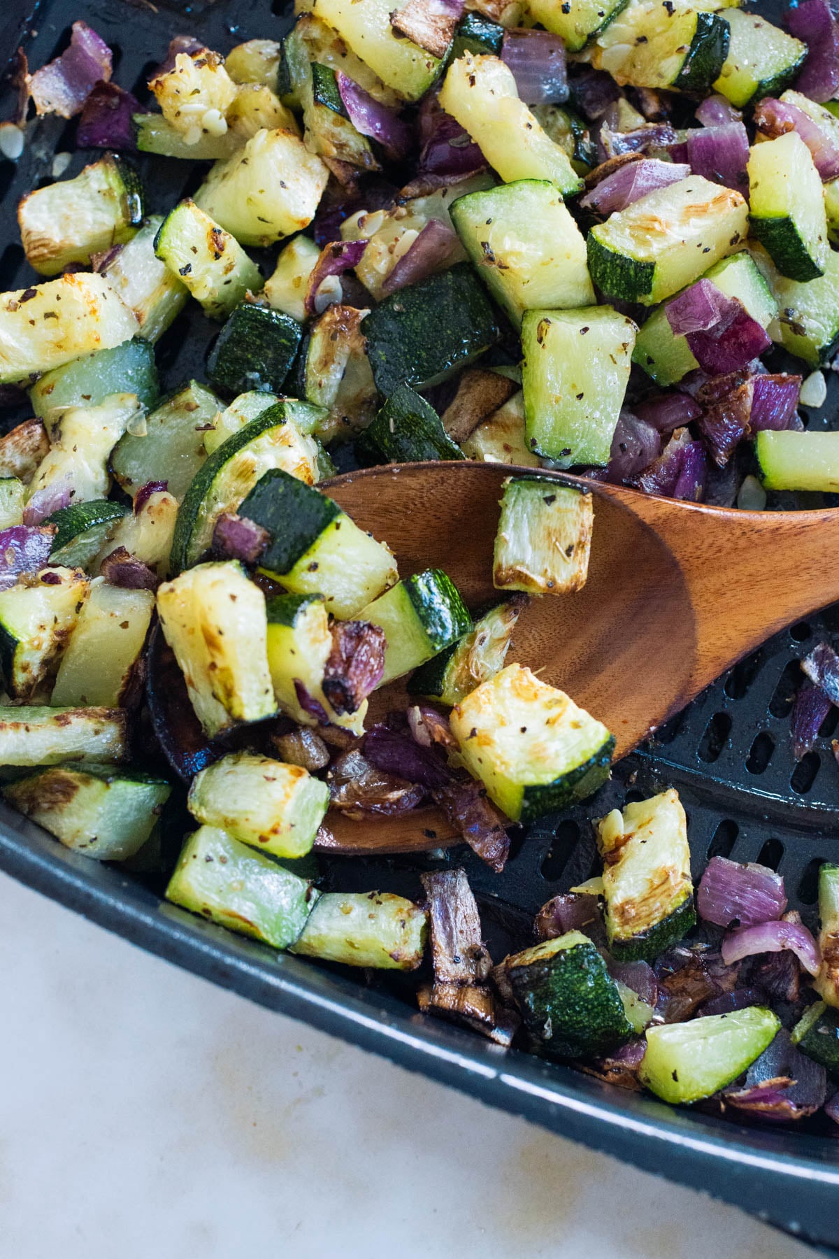 Chopped zucchini and red onion pieces being stirred with a wooden spoon in an air fryer basket.