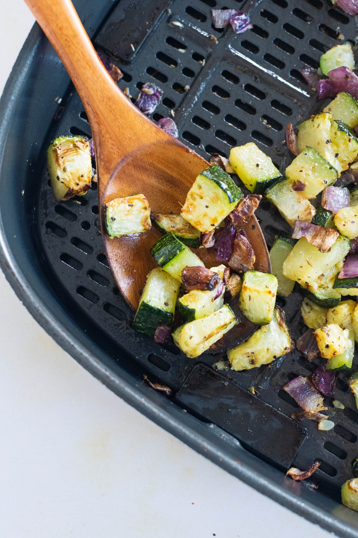 Wooden spoon scooping cooked zucchini and red onion pieces from an air fryer basket.