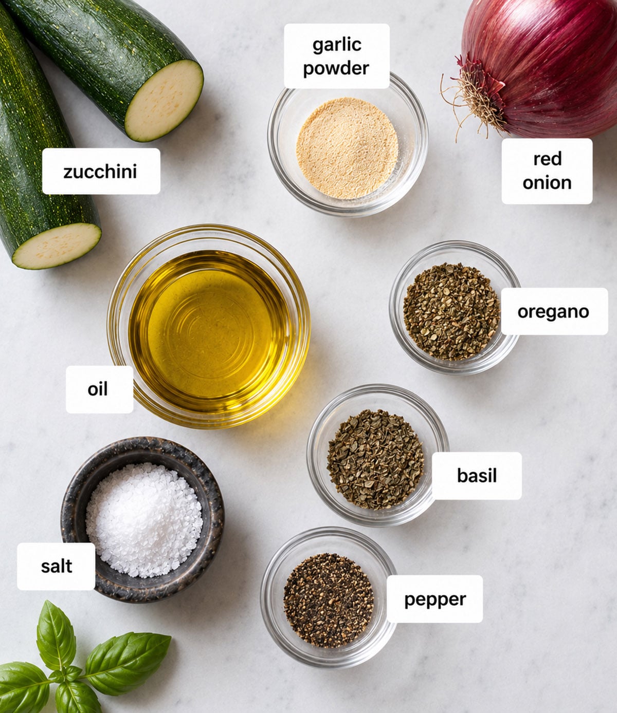 Top view of labeled ingredients on a white surface: zucchini, red onion, garlic powder, oregano, basil, pepper, oil, salt, and fresh basil leaves.