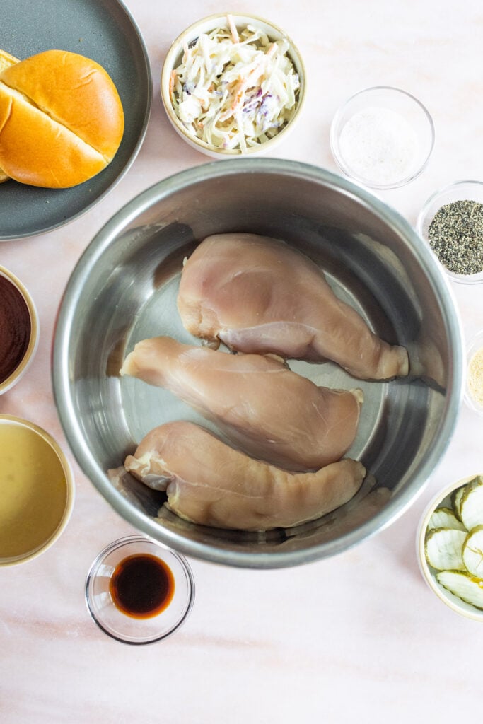 Three raw chicken breasts in a metal bowl surrounded by ingredients including sliced pickles, coleslaw, spices, a burger bun, and various liquids on a countertop.