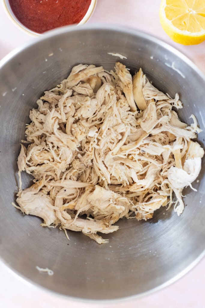 Shredded cooked chicken in a metal mixing bowl, with a bowl of red sauce and a halved lemon partially visible in the background.