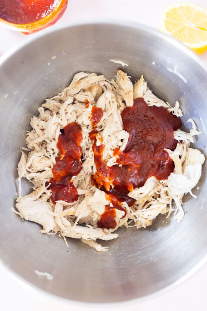 Shredded chicken in a metal bowl with barbecue sauce poured on top, next to a lemon half and a jar of sauce.