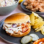 A pulled chicken sandwich with coleslaw and potato chips on a dark plate, with pickles and a bowl of barbecue sauce nearby.