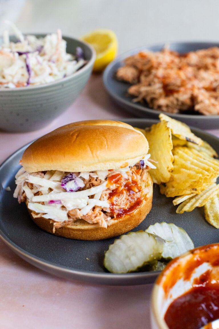 A pulled chicken sandwich with coleslaw and potato chips on a dark plate, with pickles and a bowl of barbecue sauce nearby.