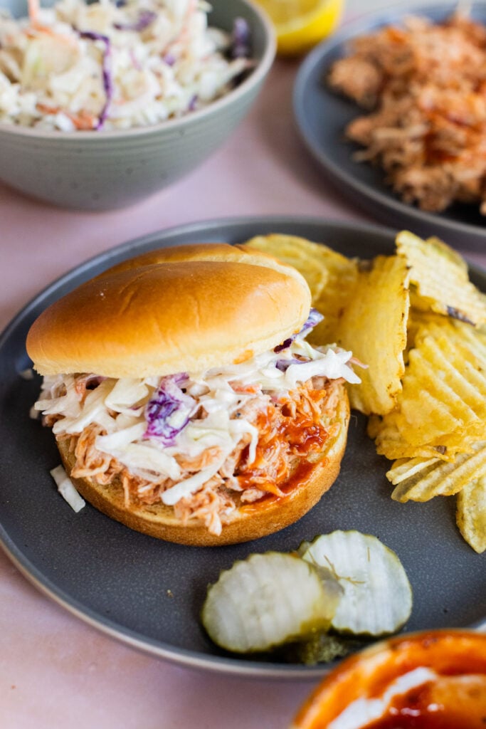 A pulled chicken sandwich topped with coleslaw, served on a gray plate with crinkle-cut potato chips and pickle slices.