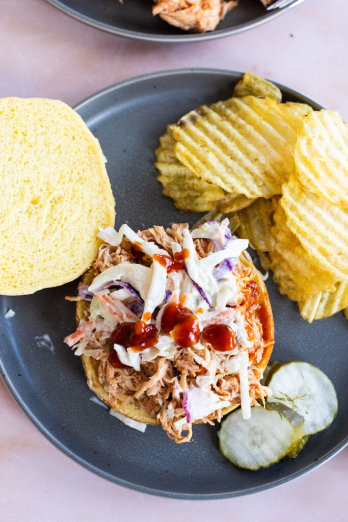 An open-faced pulled pork sandwich with coleslaw and barbecue sauce on a bun, served with crinkle-cut potato chips and pickle slices on a gray plate.