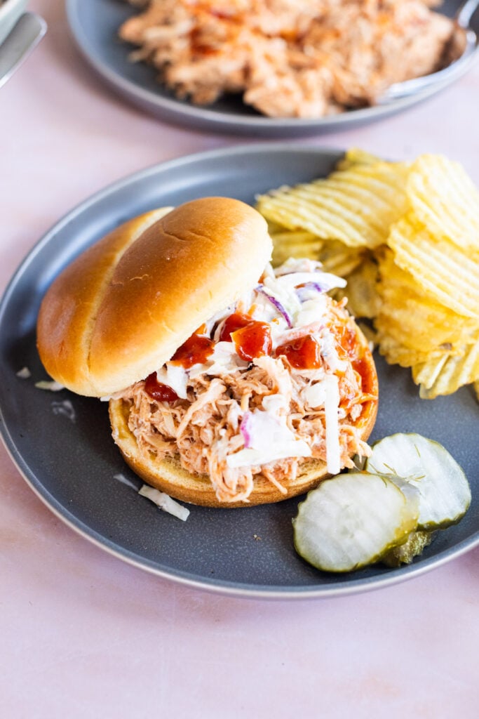 A pulled chicken sandwich with coleslaw and barbecue sauce is served on a plate with potato chips and pickle slices.
