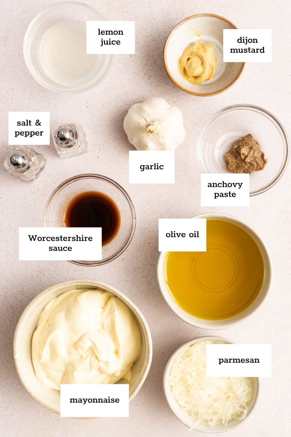A flat lay of labeled Caesar dressing ingredients: lemon juice, dijon mustard, garlic, anchovy paste, parmesan, olive oil, mayonnaise, Worcestershire sauce, and salt & pepper.