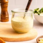 A glass jar of creamy Caesar salad dressing with a spoon inside sits on a wooden board; a pepper grinder and a bowl of salad are in the background.