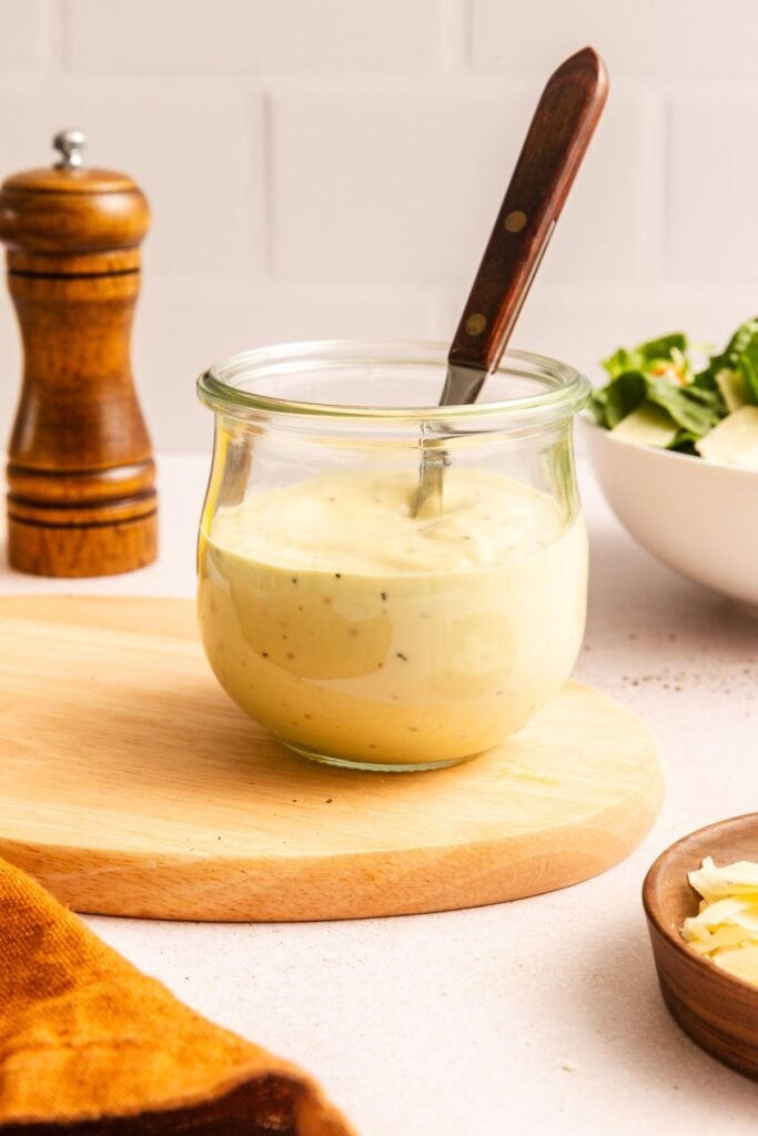 A glass jar of creamy Caesar salad dressing with a spoon inside sits on a wooden board; a pepper grinder and a bowl of salad are in the background.